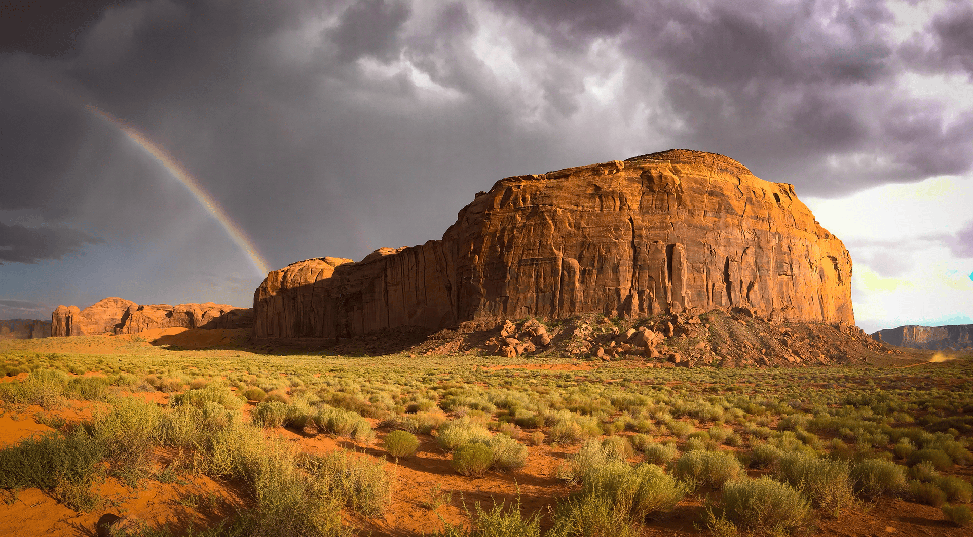 a dramatic landscape in monument valley showing a massive red sandstone butte illuminated by golden sunlight under stormy gray clouds. a rainbow arcs across the turbulent sky on the left side. the desert floor is covered with green sagebrush and scattered red rocks, creating a striking contrast between the warm earth tones and the moody sky above. 